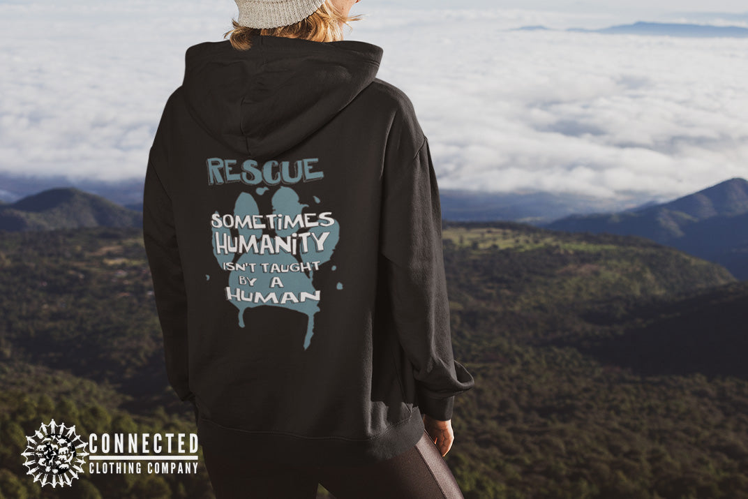 Model wearing Black Show Humanity Unisex Hoodie at the mountains that reads "Rescue. Sometimes humanity isn't taught by a human" - sharonkornman - Ethically and Sustainably Made - 10% donated to the Society for the Prevention of Cruelty to Animals